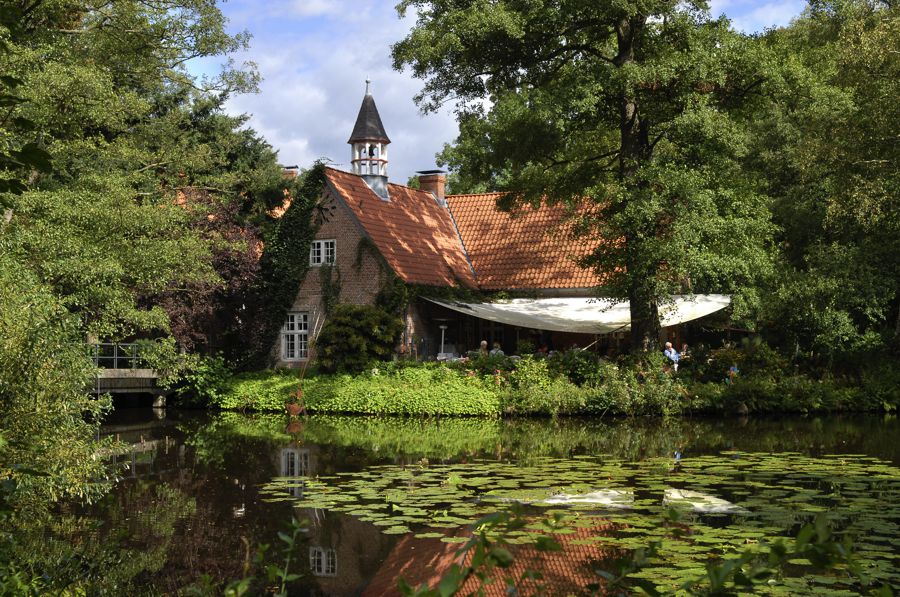 Haus an einem Teich der mit Seerosenblättern bepflanzt ist. Am Haus eine Terasse mit einigen Besuchern