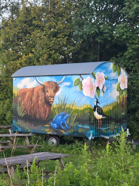 Bunt bemalter Bauwagen am Rastplatz mit Hochlandrind, blauem Frosch und Vogel vor Waldkulisse.