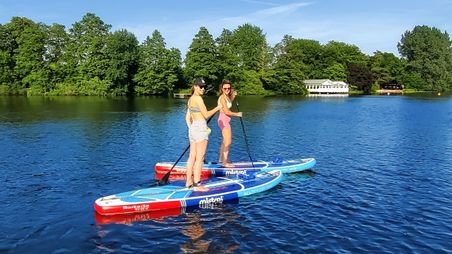 Zwei Frauen paddeln auf einem ruhigen See. Sie stehen auf bunten Paddleboards, umgeben von grünen Bäumen und einem Gebäude am Ufer.