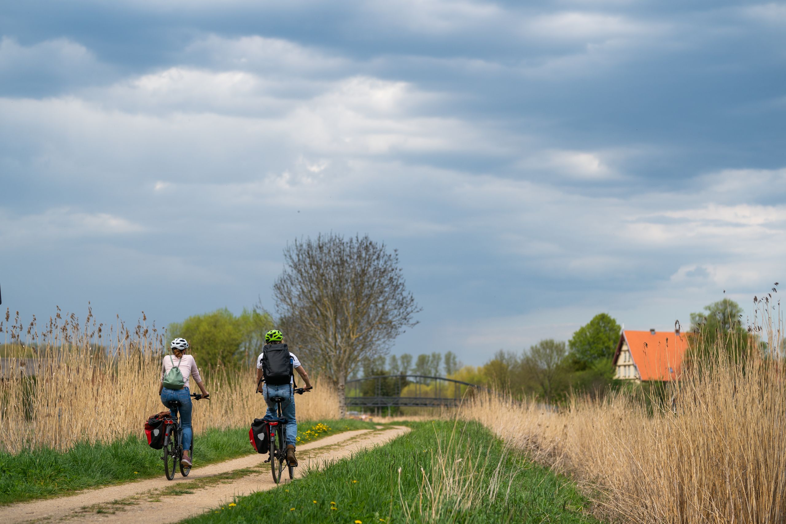 Zwei Radfahrer fahren auf einem ländlichen Weg entlang, umgeben von hohen Gräsern. Im Hintergrund ist ein Haus mit rotem Dach zu sehen.