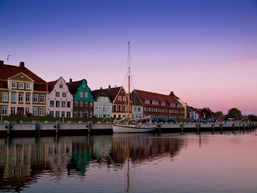 Häuser am Hafen bei Abenddämmerung, mit einem Segelboot im Vordergrund und einem klaren, farbigen Himmel im Hintergrund.