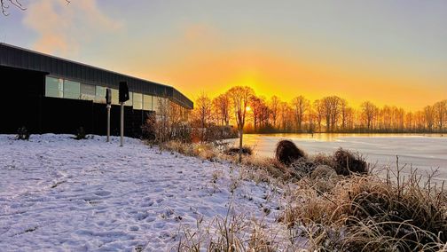 Verschneite Landschaft am Rantzauer See mit einem Schwimmbad im Vordergrund. Die Sonne geht hinter Bäumen auf und taucht die Szene in warmes Licht.