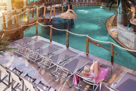 Tropisches Freizeitbad mit türkisfarbenem Innenpool, Liegen und Wasserfall; eine Person entspannt auf einer Sonnenliege.