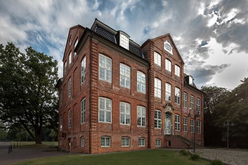 Historisches Backsteingebäude mit Treppe und Parkanlage unter wolkigem Himmel.