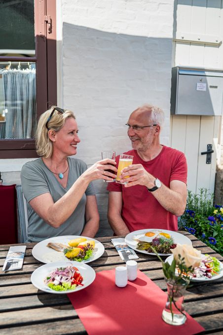 Paar stößt im Freien in Glückstadt an, vor ihnen stehen Teller mit Fischgerichten und Salat.