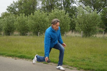 Beim Bosseln wirft ein Mann in blauer Jacke auf einem Feldweg; im Hintergrund Wiese, Zaun und Bäume.