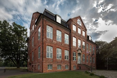 Historisches Backsteingebäude mit Treppe und Parkanlage unter wolkigem Himmel.