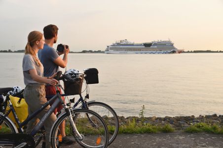 Zwei Personen mit Fahrrädern stehen am Ufer der Elbe und beobachten ein vorbeifahrendes Kreuzfahrtschiff bei Sonnenuntergang.