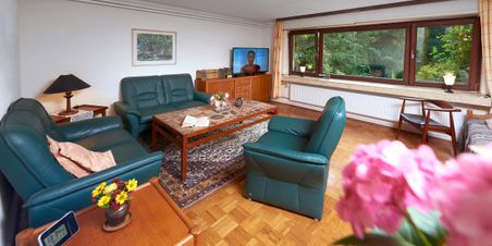Gemütliches Wohnzimmer mit grünen Ledermöbeln, Couchtisch, Fernseher und großem Fenster mit Blick in den Garten.