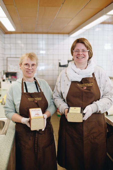 Zwei Frauen in braunen Schürzen präsentieren Käselaibe in einer gefliesten Küche. Sie lächeln und tragen Handschuhe.