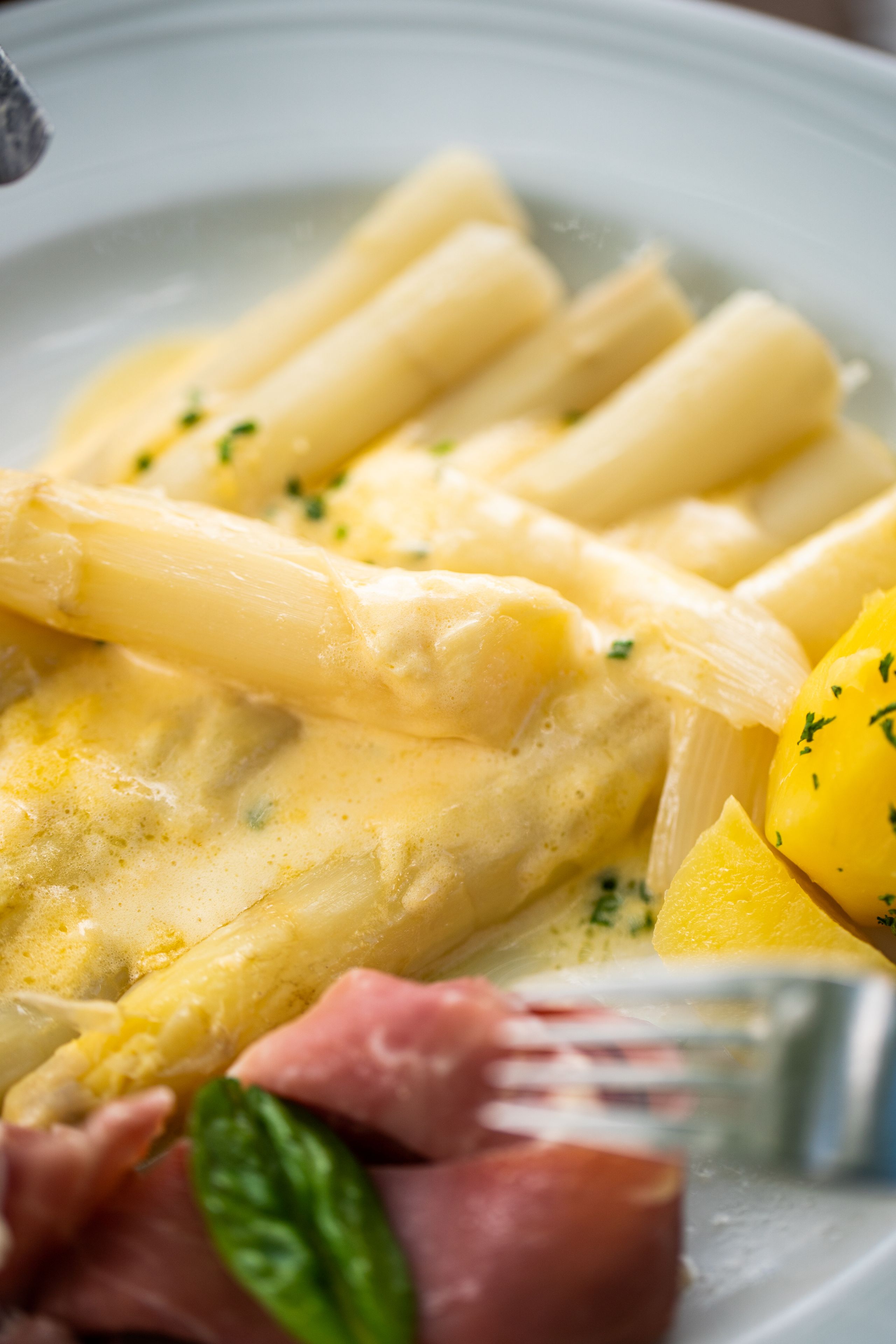 Spargel mit Sauce Hollandaise, serviert mit Kartoffeln und Schinken. Ein klassisches Gericht, das auf einem weißen Teller angerichtet ist.