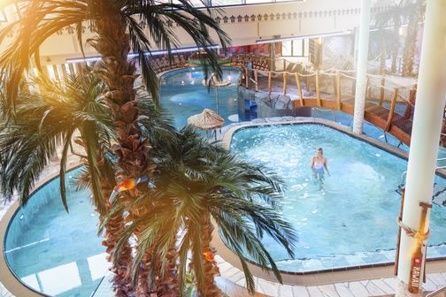 Tropisch gestaltetes Freizeitbad mit Innenpool, Palmen und Holzbrücke; eine Frau steht im Wasser.