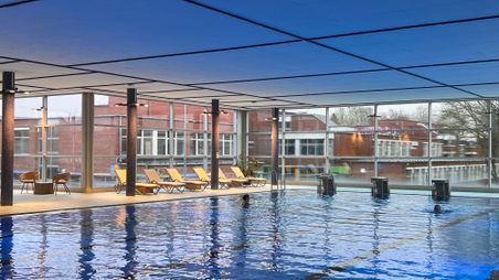 Ein modernes Hallenbad mit großen Fenstern, Schwimmern im Wasser und Liegestühlen am Beckenrand. 
