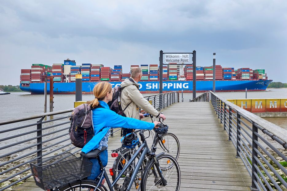 Ein Paar bewegt sich mit den Fahrrädern schiebend in Richtung Willkomm-Höft und blickt auf ein Containerschiff auf der Elbe.