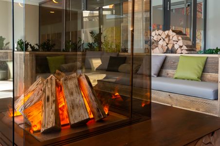 Ein moderner Kamin in einem Lounge-Bereich mit Holzscheiten, Sitzbänken und Kissen. Die Umgebung wirkt einladend und gemütlich.