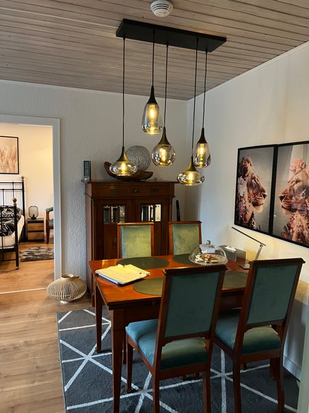 Gemütlicher Essbereich mit Holztisch, grünen Stühlen, fünf Pendelleuchten und Tierbildern an der Wand; links Blick ins Schlafzimmer.