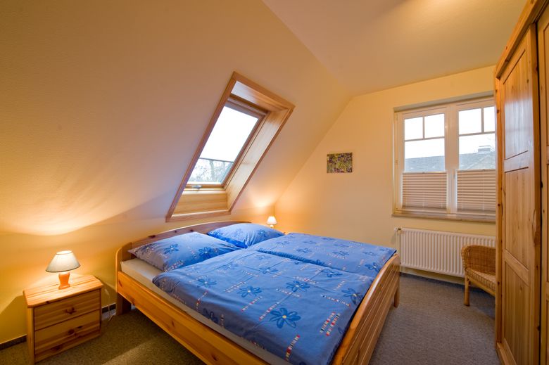 Ein gemütliches Schlafzimmer mit Dachfenster, Holzmöbeln und blauem Bettzeug. Eine kleine Lampe steht auf einem Nachttisch.