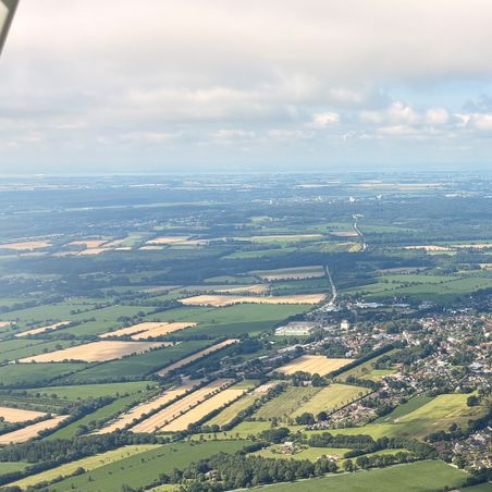 Luftaufnahme einer weiten Agrarlandschaft mit Feldern, Waldstreifen und einem Dorf unter bewölktem Himmel.