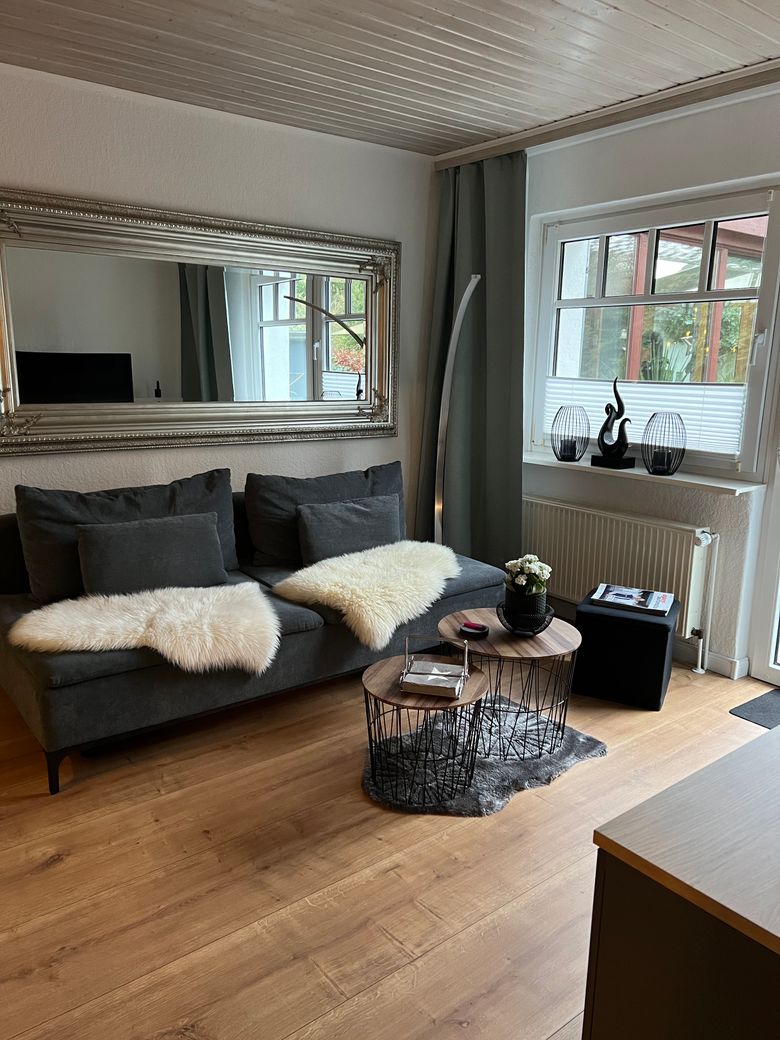 Gemütliches Wohnzimmer mit grauem Sofa, Felldecken, großem Spiegel und dekorativen Beistelltischen am Fenster.