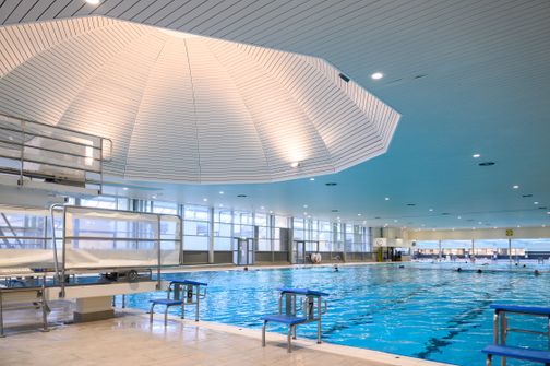 Modernes Schwimmzentrum mit großem Pool, Sprungturm und heller Beleuchtung. Die Decke hat ein auffälliges Design.