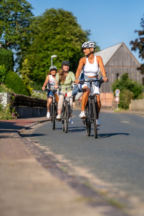 Drei Frauen fahren bei Sonnenschein mit dem Fahrrad durch eine Ortschaft; im Hintergrund stehen ein Haus und grüne Bäume.