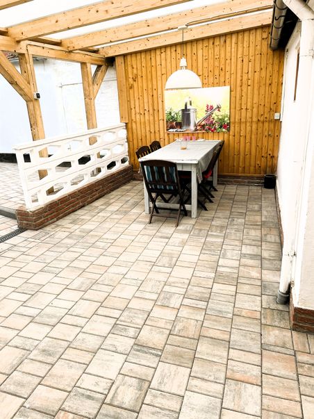 Ein überdachter Innenhof mit Holzwänden, einem langen Tisch und Stühlen. An der Wand hängt ein Bild mit Gartenmotiv. Der Boden ist gefliest.
