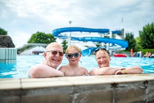 Eine Familie genießt einen Tag im Schwimmbad. Im Hintergrund ist eine große blaue Wasserrutsche zu sehen. Alle lächeln und tragen Schwimmbrillen.
