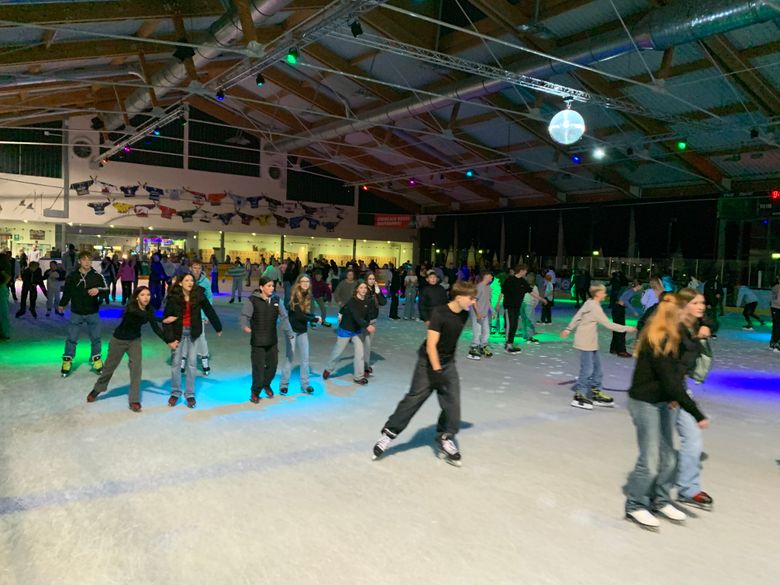 Viele Menschen auf Schlittschuhen in einer beleuchteten Eishalle mit Disco-Kugel und bunten Lichtakzenten.