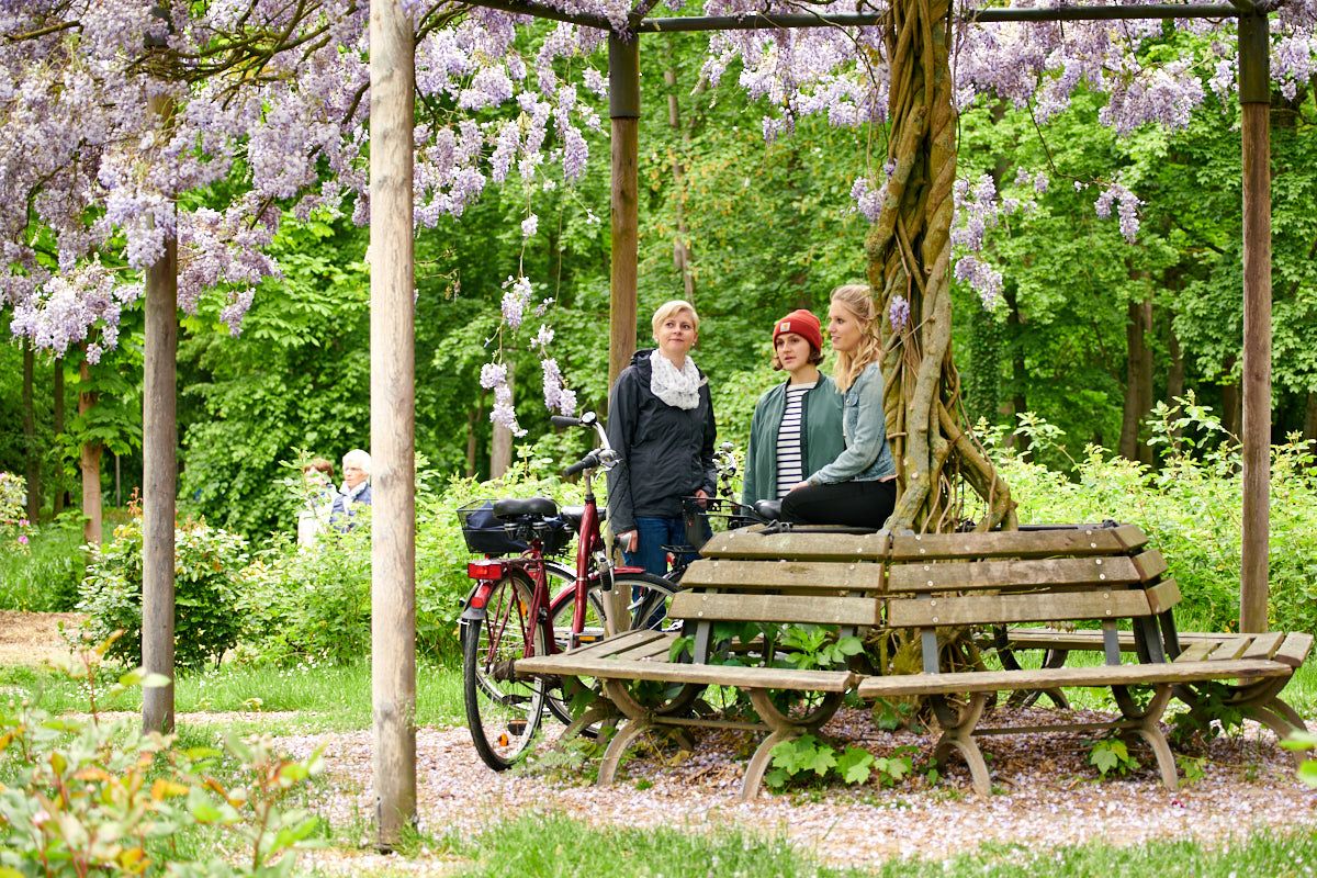 Drei Personen entspannen unter einer blühenden Pergola im Park. Fahrräder lehnen an der Bank, umgeben von üppigem Grün und lila Blüten.