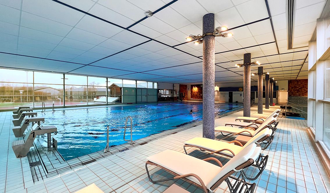 Ein modernes Hallenbad mit einem großen Schwimmbecken, umgeben von Liegestühlen. Große Fenster lassen viel Licht herein.
