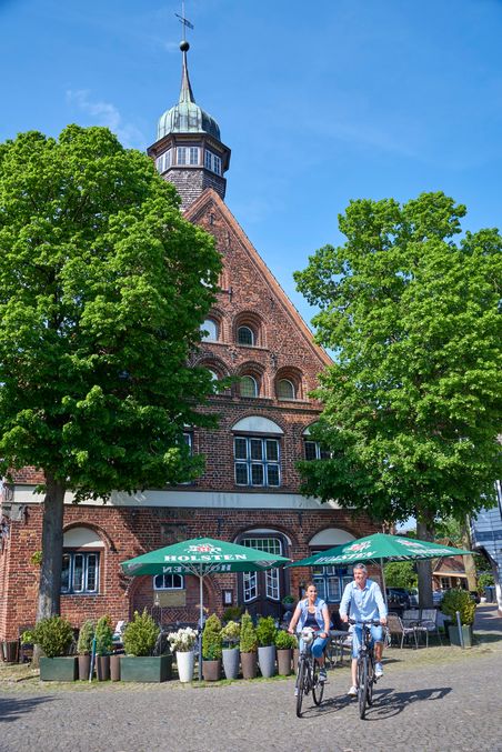 Zwei Radfahrer vor einem historischen Backsteingebäude mit Turm und grünen Bäumen. Im Vordergrund sind Holsten-Sonnenschirme zu sehen.