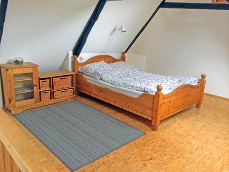 Gemütliches Dachgeschosszimmer mit Holzbett, Schrank mit Körben und grauem Teppich auf Holzboden. Weiße Wände und sichtbare Dachbalken.