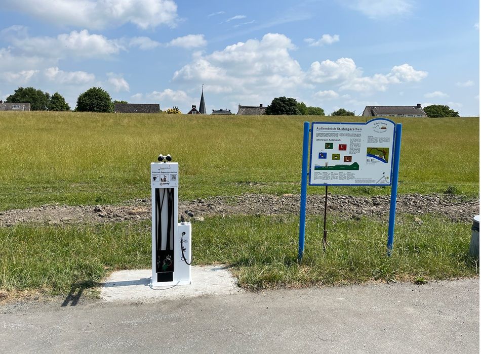 Wiese mit Schild „Außengelände St. Margarethen“ und Fahrrad-Reparaturstation, dahinter Häuser und Kirchturm unter blauem Himmel.