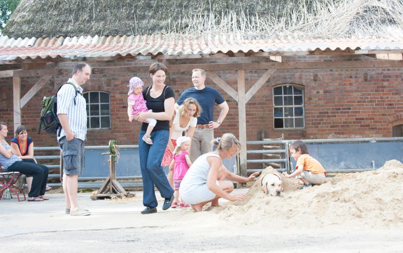 Mehrere Erwachsene und Kinder auf dem Hof Schürmann; ein Kind spielt im Sand mit einem Hund vor einem Backsteingebäude.