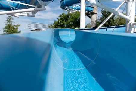 Blaue Wasserrutsche im Freibad unter blauem Himmel. Die Rutsche schlängelt sich durch die Anlage, umgeben von Bäumen.