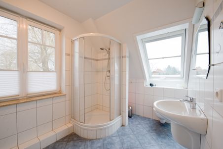 Modernes Badezimmer mit Eckdusche, Waschbecken und zwei Fenstern. Die Wände sind weiß gefliest, der Boden hat blaue Fliesen.