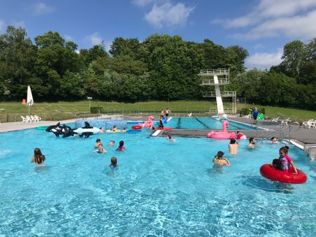 Kinder planschen in einem Freibad mit aufblasbaren Tieren und Schwimmringen. Im Hintergrund ist ein Sprungturm zu sehen.