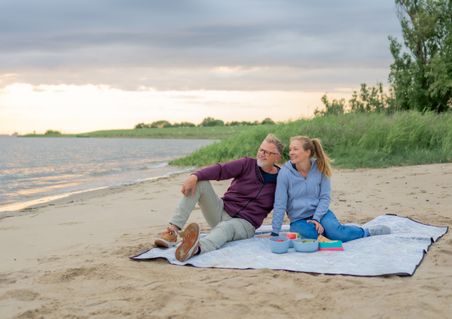 Ein Paar sitzt auf einer Decke am Elbstrand und genießt ein Picknick bei Sonnenuntergang. Der Himmel ist bewölkt, und das Wasser glitzert im Abendlicht.