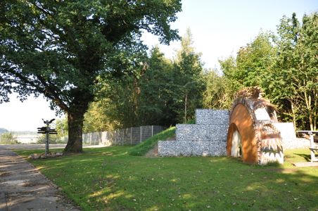 Ein grüner Aussichtspunkt mit einem großen Baum, einem Wegweiser und einer Skulptur aus Metall und Steinen. Der Bereich ist von Bäumen umgeben.