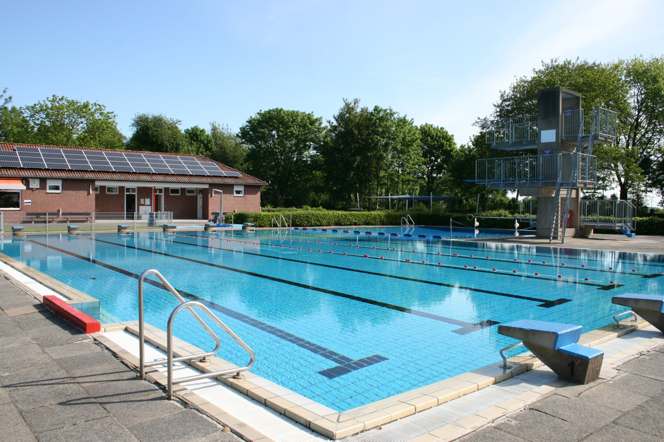 Freibad mit einem großen Schwimmbecken, Sprungturm und einem Gebäude mit Solarpanelen auf dem Dach. Der Himmel ist klar und sonnig.