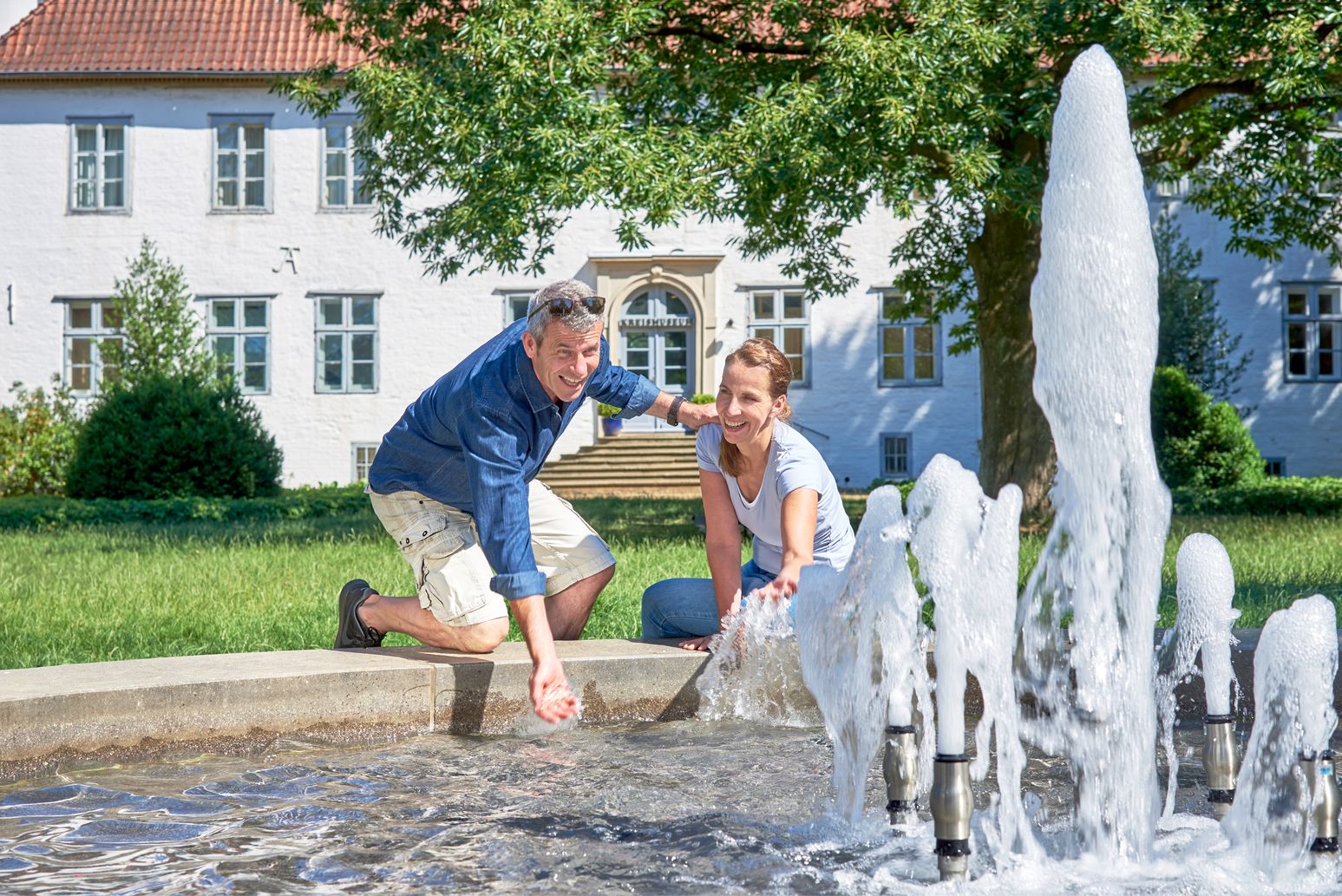 Zwei Personen spielen am Springbrunnen vor dem Kreismuseum Itzehoe in Holstein; im Hintergrund steht das weiße Museumsgebäude.