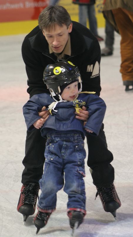 Ein Mann hilft einem kleinen Kind beim Eislaufen in einer Eishalle. Beide tragen Schlittschuhe, das Kind zusätzlich einen Helm.