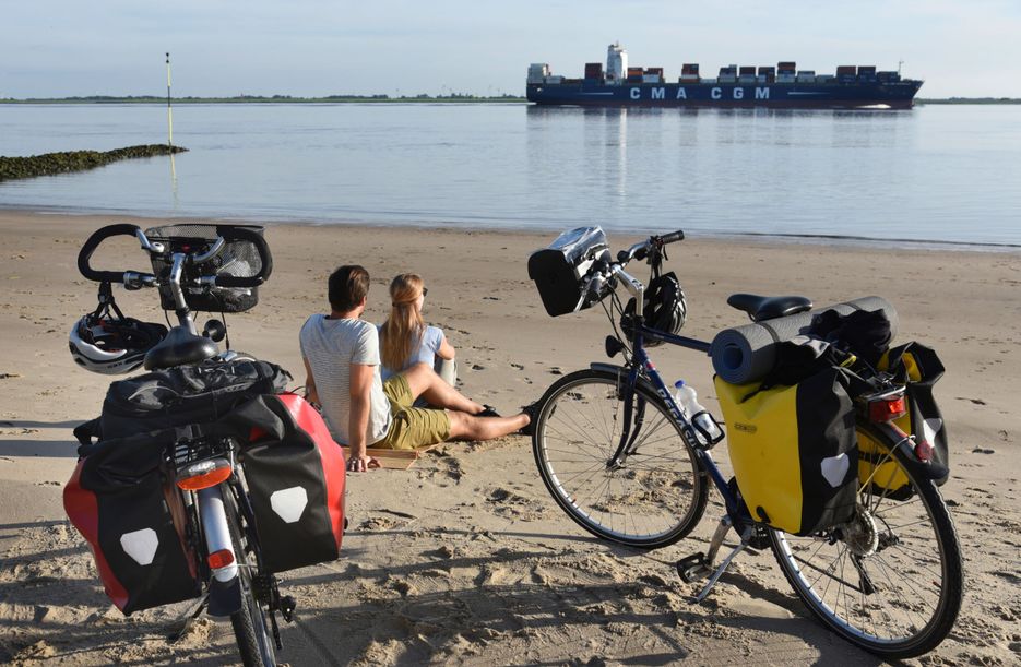 Paar ist mit dem Fahrrad auf dem Elberadweg unterwegs und genießt eine Pause am Elbstrand in Brokdorf und beobachtet die Schiffe.