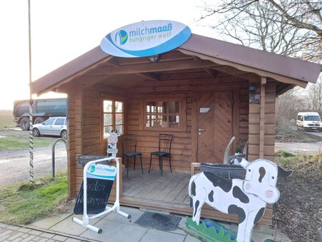 Ein kleines Holzhaus mit einem Schild "milchmaab hungriger wolf". Davor steht eine bemalte Kuhfigur und ein Willkommensschild.