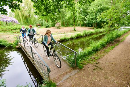 Drei Personen fahren mit Fahrrädern über eine kleine Brücke in einem grünen Park. Der Weg führt entlang eines Kanals, umgeben von Bäumen und Blumen.