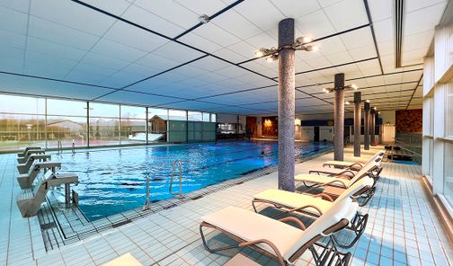 Ein modernes Hallenbad mit einem großen Schwimmbecken, umgeben von Liegestühlen. Große Fenster lassen viel Licht herein.