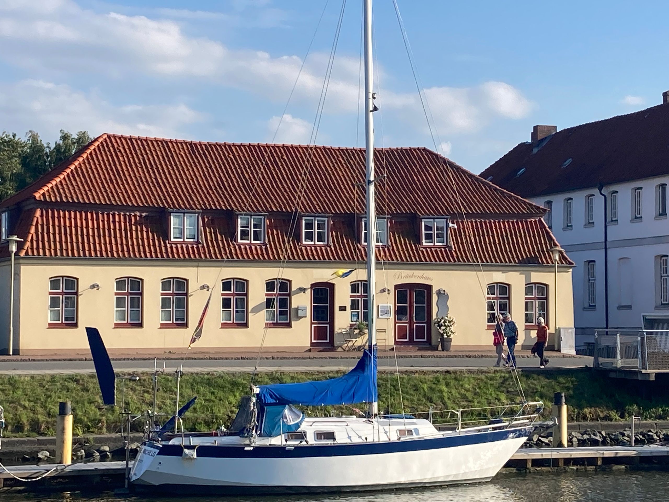 Das Brückenhaus am Hafen mit rotem Ziegeldach, Segelboot im Vordergrund und Spaziergängern am Steg.
