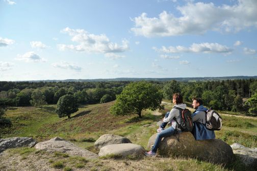 Zwei Wanderer sitzen auf Felsen und genießen den Blick über die grüne Hügellandschaft unter blauem Himmel.