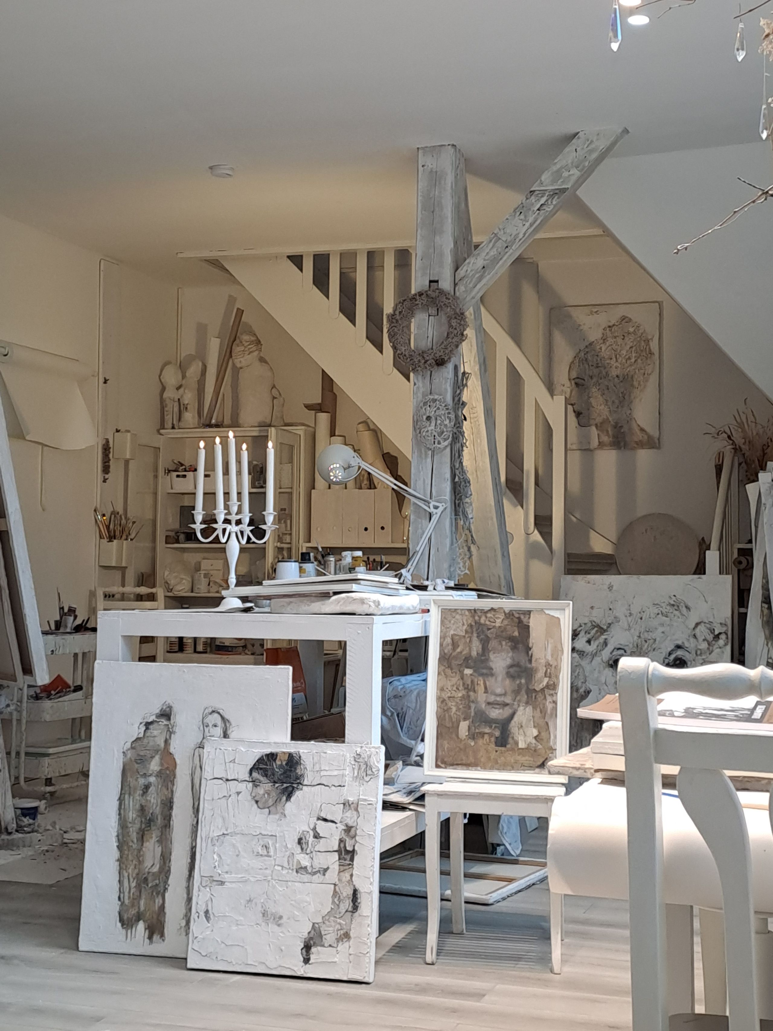Helles Atelier mit weißen Möbeln, Staffeleien und abstrakten Porträtgemälden; im Hintergrund Treppe, Kerzenständer und Kunstobjekte.