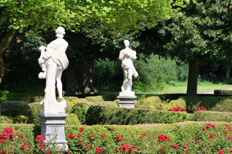 Zwei weiße Statuen stehen in einem gepflegten Garten, umgeben von roten Rosen und grünen Hecken. Im Hintergrund sind Bäume zu sehen.